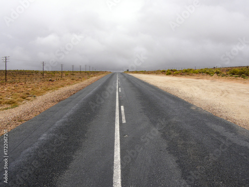 Bild auf Leinwand Carretera asfaltada que se pierde en el horizonte entre terrenos áridos, con postes telefónicos a un lado y un cielo gris cargado de nubes