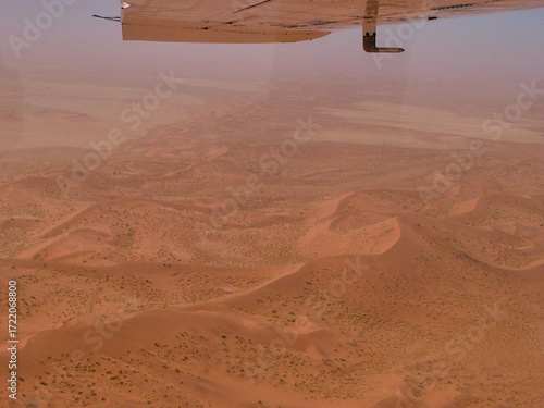 Konstfotografi Vista aérea de un desierto de dunas rojizas con relieves ondulados, captado desde un avión cuya ala aparece en la parte superior, bajo un cielo claro