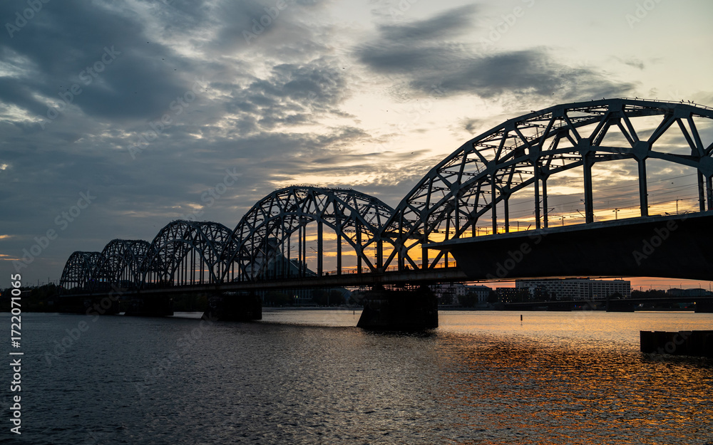 Naklejka premium A railway bridge spanning over a river at sunset, with warm golden light reflecting on the water. 