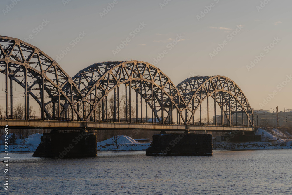 Fototapeta premium Sunset landscape of an iron-framed arched bridge crossing a river in a snowy winter