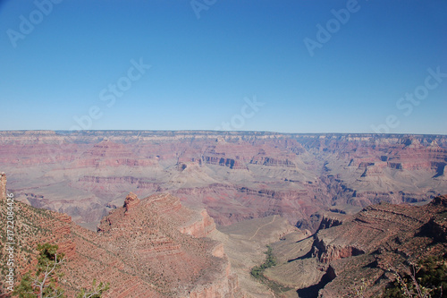 Panoramic view of the Grand Canyon with layered red rock formations, steep cliffs, and wide valleys beneath a clear blue desert sky