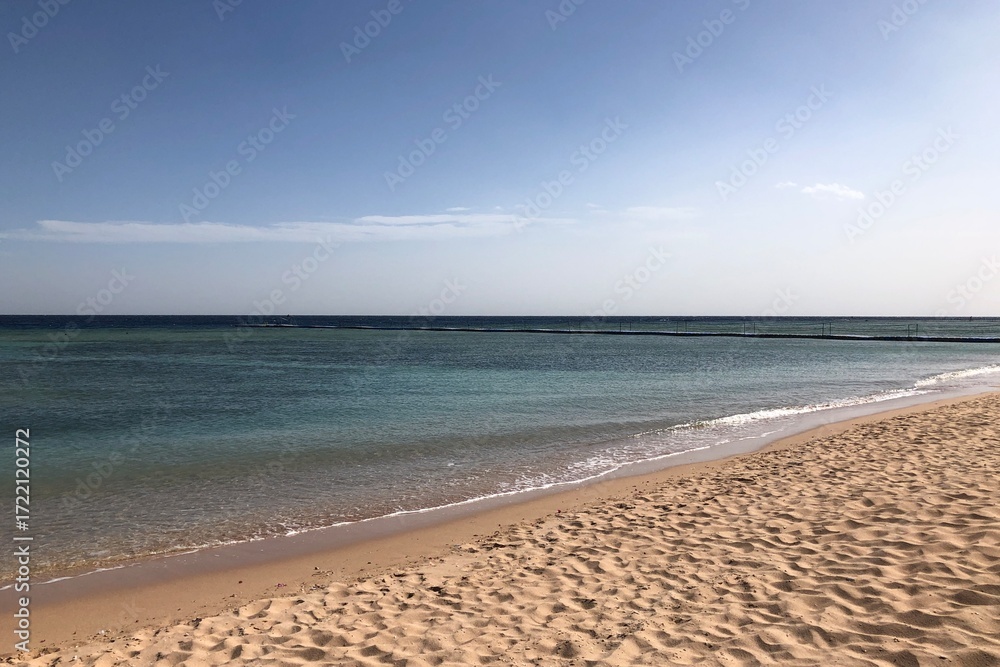 Fototapeta premium Turquoise water gently meeting a sandy beach under a clear sky. A peaceful seaside scene with gentle waves lapping the shore under the summer sun