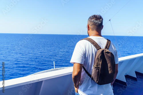 Young man with backpack standing on the deck of a ferry boat, admiring the breathtaking view of the mediterranean sea while traveling to the balearic islands during summer holidays.
