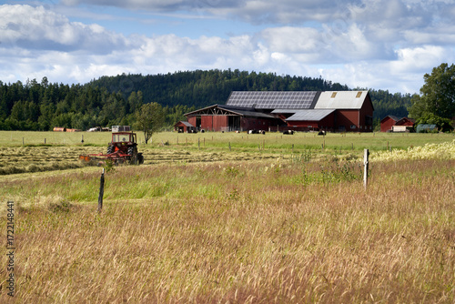  Red Tractor Harvesting Hay in Rural Scandinavian Landscape with Traditional Farm Buildings, Dollar Panels, and Countryside Backdrop Near Håverud, Sweden