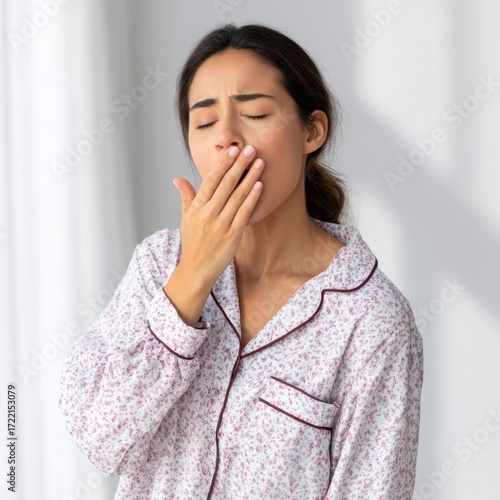 A tired young woman in patterned pajamas yawns with her eyes closed, covering her mouth with her hand, suggesting morning grogginess or lack of sleep.