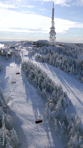 Aerial view of snowy mountain resort with ski lifts, forest, and tall tower on the summit. Perfect for themes of winter, sports, travel, tourism, and scenic alpine landscapes.