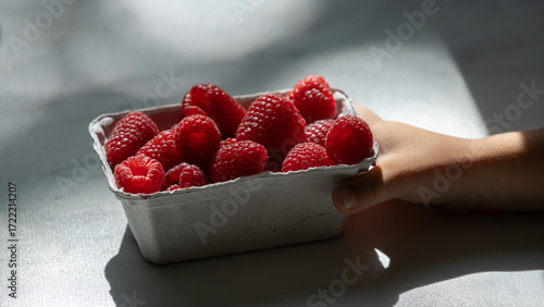 Little hand holding carton of fresh raspberries in natural light