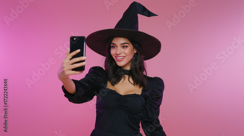 Young woman wearing a black witch costume with hat smiling while taking a selfie with smartphone on pink background celebrating halloween