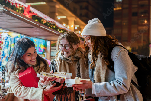 Women friends laughing while sharing food and shopping at a winter christmas market