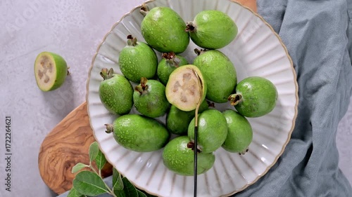 A collection of freshly picked feijoa fruits, some with glistening water droplets, are arranged on a textured white plate, a branch of leaves laying on the side.