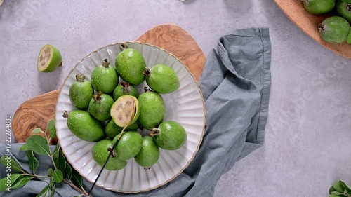 A collection of freshly picked feijoa fruits, some with glistening water droplets, are arranged on a textured white plate, a branch of leaves laying on the side.