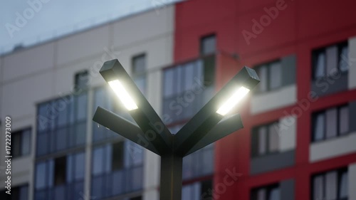 Three-armed LED streetlight glowing at dusk in front of a modern residential facade with red panels, glass balconies and windows, emphasizing contemporary urban architecture and geometric lines