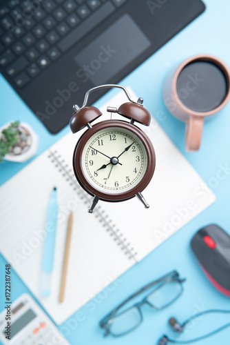 Maximize Productivity with This Streamlined Workspace, Featuring a Classic Alarm Clock, Laptop, and Coffee on a Light Blue Surface for a Focused Work Session