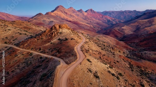 An aerial view of a winding dirt road traversing a mountainous landscape, showcasing diverse earthy tones and textures at sunrise or sunset
