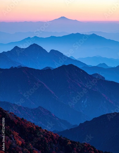 Layered blue mountains recede into a soft pink and blue dawn. Valleys filled with trees in autumn colors sit in the foreground