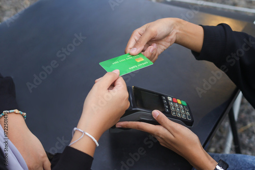 Credit card payment using a point of sale terminal. Close-up of hand swiping a green card while another person waits at a table. Modern cashless transaction concept.