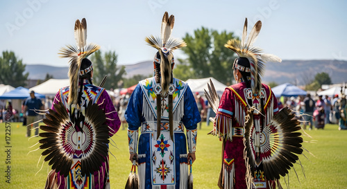 Three Native American men in traditional regalia and headdresses standing on a grassy field during an Indigenous Peoples Day celebration with a clear sky and distant mountains.