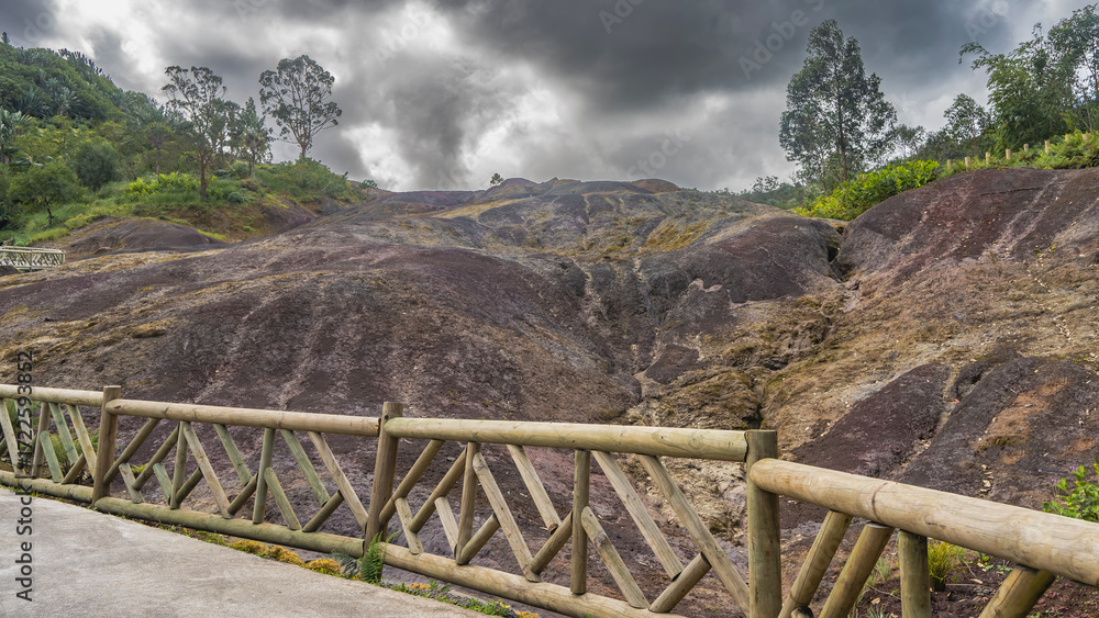 Fototapeta premium A unique natural landmark, 23 coloured earth. Colorful sand hills. Green vegetation on the mountainside. Wooden railings along the footpath. Clouds in the sky. Mauritius. La Vallée Des Couleurs
