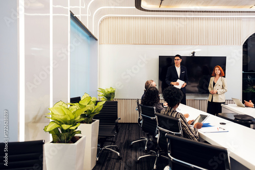 Modern office boardroom where a team watches presenters by a large screen; sleek interior with plants and black chairs, capturing corporate collaboration, strategy, and training.