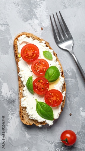Toast with creamy spread, halved cherry tomatoes, and basil leaves, next to a fork on a gray, textured surface