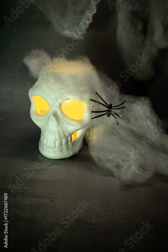 Skull with glowing yellow eyes sits amid cobwebs and a black spider, casting warm light against a dark background. A classic Halloween setup creating a spooky, eerie, and festive atmosphere.