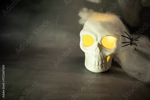 Skull with glowing yellow eyes sits amid cobwebs and a black spider, casting warm light against a dark background. A classic Halloween setup creating a spooky, eerie, and festive atmosphere.