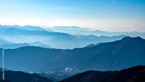 Mountains fade into blue distance under clear sky, with a small village nestled in a valley surrounded by textured ridges