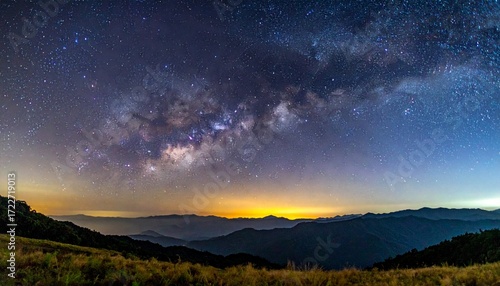 Panoramic vista of the Milky Way galaxy over rolling mountains. Soft golden light radiates on the horizon. Forested hills fade into the distance