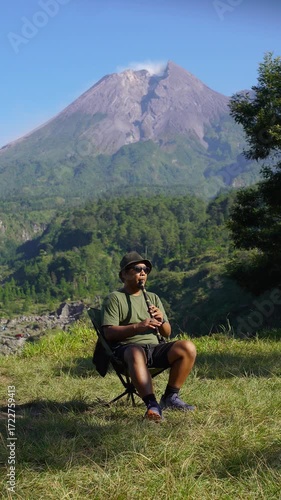 A solo male adventurer plays a traditional instrument called a low whistle against a backdrop of a volcano. A solo camper plays a low whistle on a bright morning against a backdrop of a volcano.