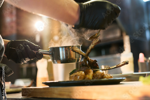 Fotografija Caucasian young adult man wearing black gloves plating cooked lamb chops and veg