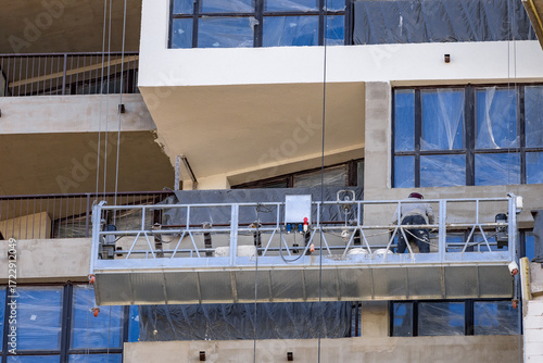 Construction worker on suspended platform performs facade plastering on tall urban building, wearing safety gear and harness, restoring exterior wall surface. Worker applies plaster on building facade