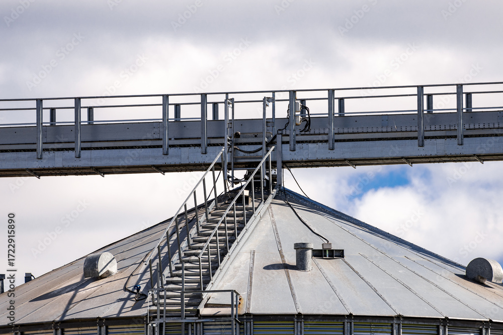 Fototapeta premium A high-angle shot shows several large, silver grain silos under a partly cloudy blue sky. Metal walkways and structures connect the silos. The scene suggests an industrial agricultural facility.
