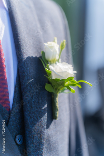 A man in a gray jacket and white shirt. It has a black bowtie on it. In the jacket pocket there is a bouquet of white flowers with green leaves.