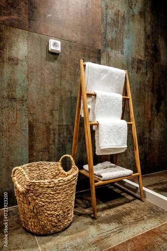 Bathroom corner with wooden ladder holding towels and wicker laundry basket against rustic tiled wall. Modern spa-style interior design combining functionality, warmth, and natural elements.