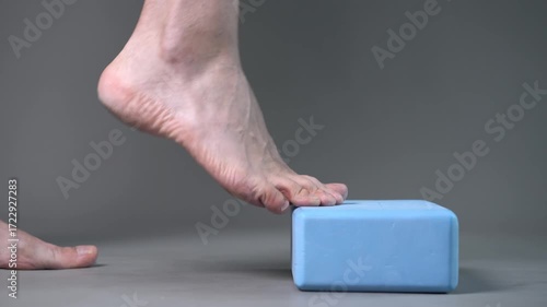 Individual balance training on a blue block with bare feet, demonstrating foot position and stability on a gray studio background