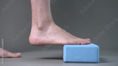 Individual balance training on a blue block with bare feet, demonstrating foot position and stability on a gray studio background