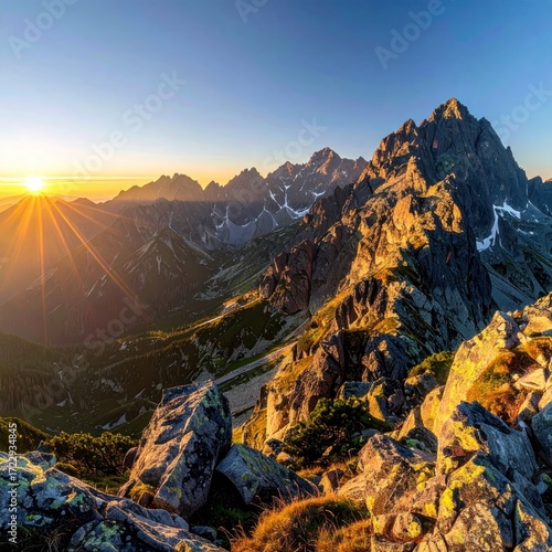 Sun illuminates rocky mountain peaks, casting golden light across the ridges. Vegetation covers the slopes amidst a clear blue sky backdrop