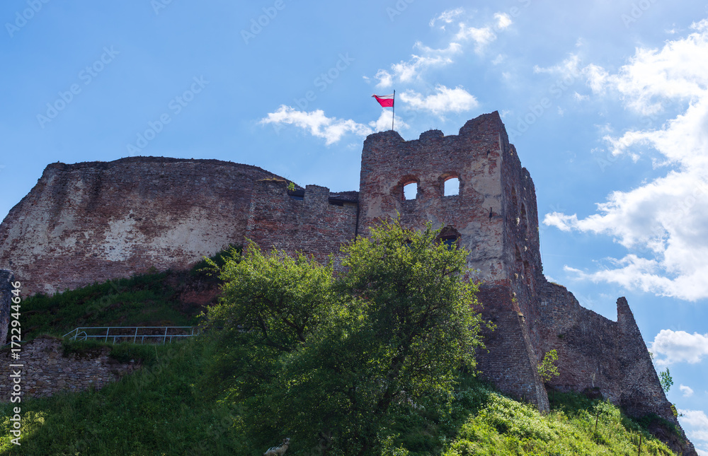 Fototapeta premium The stone ruins of a medieval castle tower rise against a blue summer sky, with the Polish national flag waving proudly on top. A historical landmark symbolizing cultural heritage and centuries of his