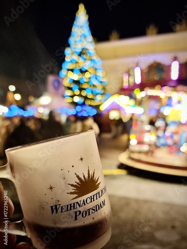  Christmas market scene in Potsdam, Germany. Large Christmas tree adorned with blue and gold lights Hand holds a festive mug inscribed with 
