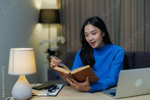 фотографія Young Asian woman sitting at her desk, engrossed in a book under the warm light