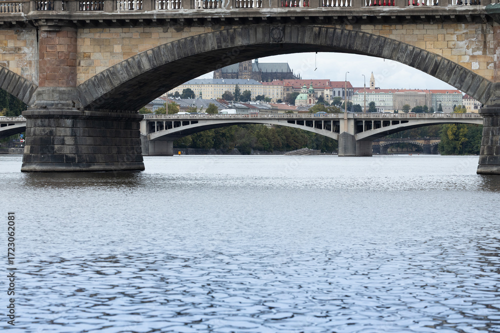 Naklejka premium Architectural view of historic stone bridges spanning a river, with reflections on the water and a backdrop of city buildings under a cloudy sky showcasing urban beauty