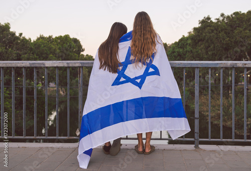 Two Israeli teen girls wrapped in the flag of Israel, looking over a bridge