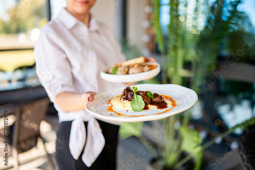Restaurant server holding two plated meals with meat, mashed potatoes, and vegetables in a bright, upscale dining setting. Food presentation and hospitality concept