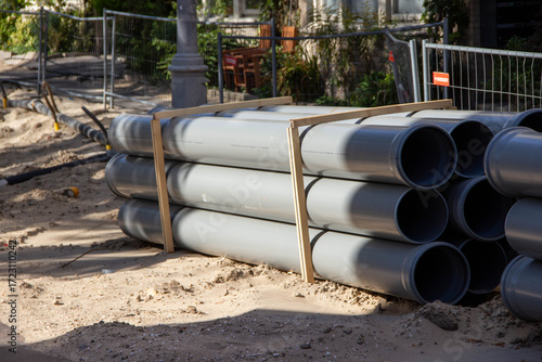 Wallpaper Mural Stacked gray PVC pipes secured with wooden supports on a construction site, surrounded by sand and fencing, illustrating materials for urban infrastructure development Torontodigital.ca