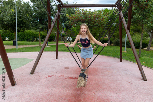 Smiling Girl Balancing on Rope Swing in Park. A happy young girl stands on a rope swing at an outdoor playground, confidently balancing and smiling in a green park setting.