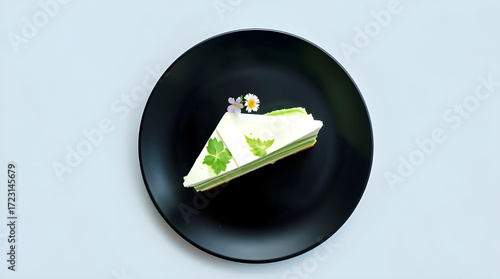Minimalist Flat Lay of a Triangular Slice of Green Layered Mousse Cake with Flower Garnish on a Black Plate and Solid Light Background