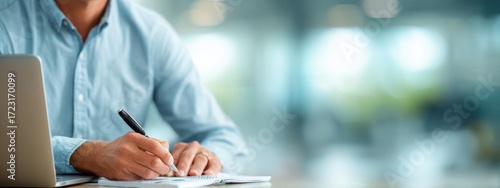 a businessman in a shirt is working on a laptop computer he taking notes or participating in a meeting in a conference room, using the laptop for a financial concept. the background is blurred.