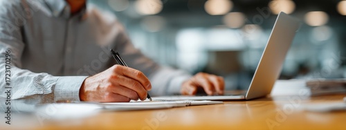a businessman in a shirt is working on a laptop computer he taking notes or participating in a meeting in a conference room, using the laptop for a financial concept. the background is blurred.