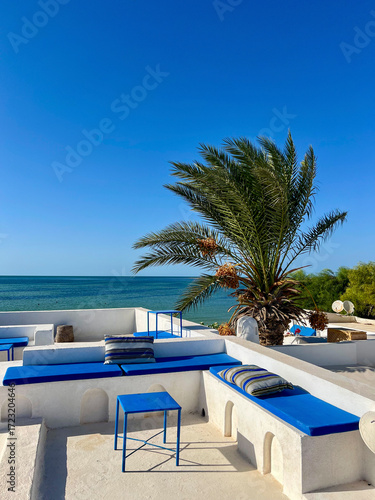 Enjoy the stunning scenery from a cozy rooftop in Djerba, Tunisia. The whitewashed terrace features blue seating, offering a perfect view of the sea and nearby palm trees.