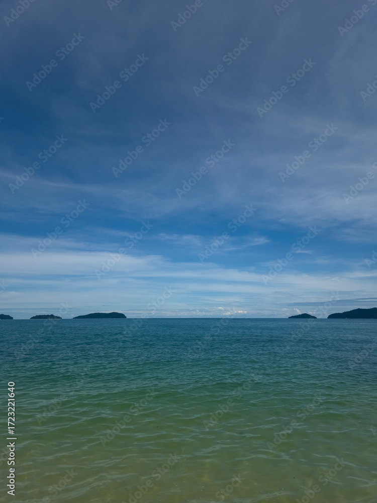 Fototapeta premium Calm tropical seascape with turquoise ocean water, blue sky and distant islands on the horizon under soft white clouds, peaceful summer vacation background in Southeast Asia beach landscape